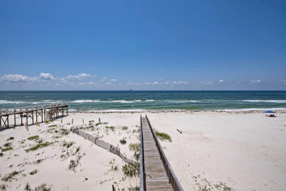 A wooden boardwalk stretches out over soft, white sand towards the gentle ocean waves. Flanked by lush, coastal grasses, the path promises direct, easy access from the property to a secluded beach area.