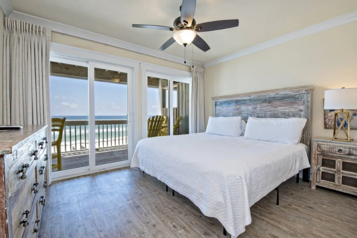 A king bedroom featuring a plush bed positioned to face a sliding glass door that opens onto a balcony with an ocean view. The room is styled with serene, neutral décor and includes a dresser and soft ambient lighting, creating a calm, luxurious retreat.