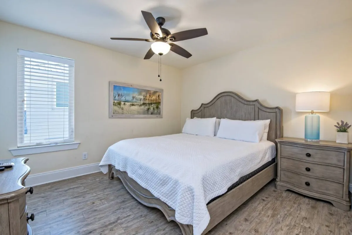 A tranquil queen or king bedroom with soft, natural light streaming through the window. The bed is dressed in plush linens, accompanied by a nightstand and tasteful artwork, exuding peace and comfort.