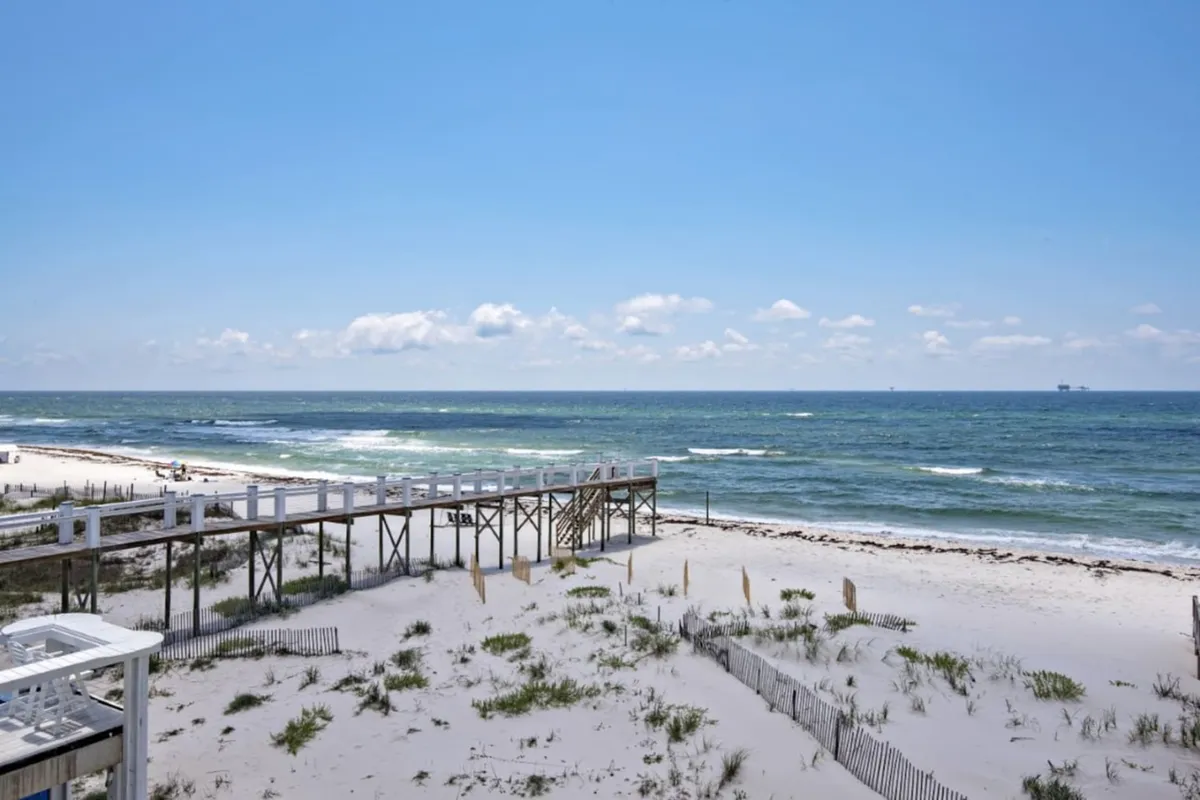 A captivating view of the sandy beach, a long pier stretching into the ocean, and waves gently rolling ashore. The scene also includes open sky and hints of beach activity, creating a relaxed and engaging coastal vibe.