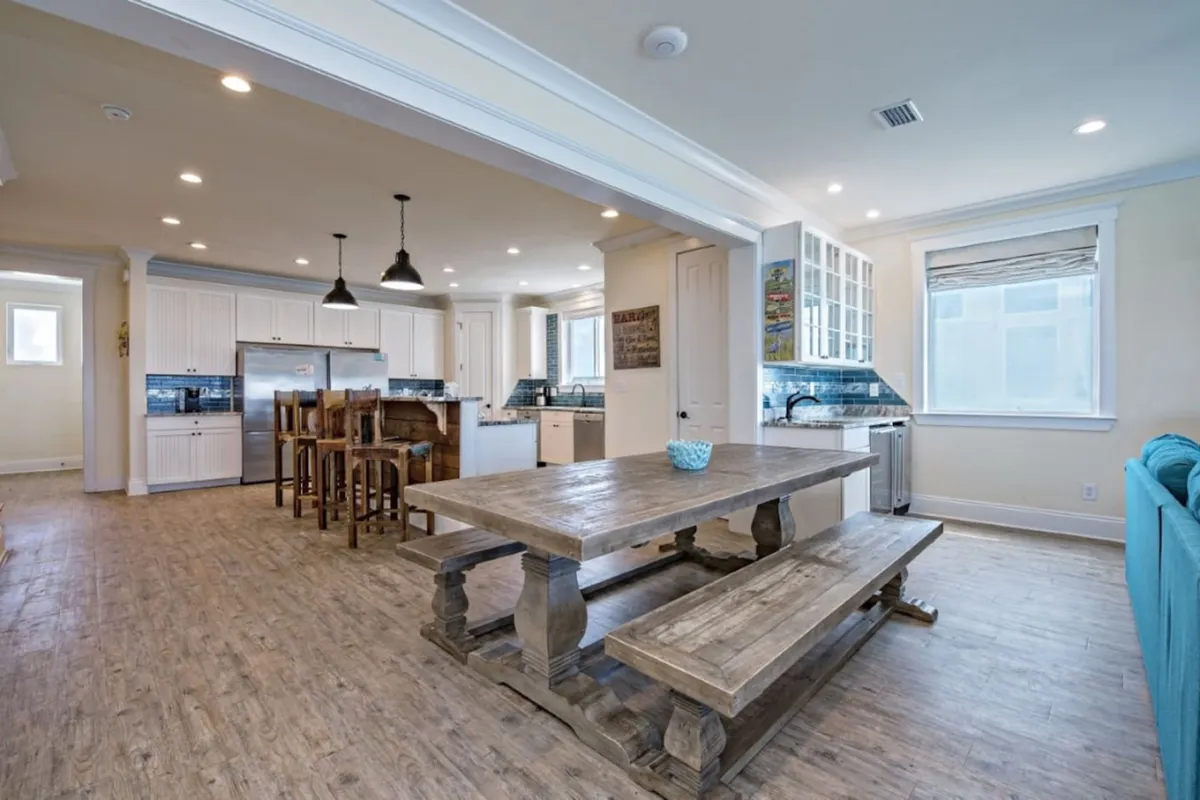 This image features a large wooden dining table adjacent to a kitchen island with bar stools and modern appliances. The open layout creates a sense of communal space, ideal for shared meals and celebrations. The design is both functional and welcoming, fostering an atmosphere of togetherness.