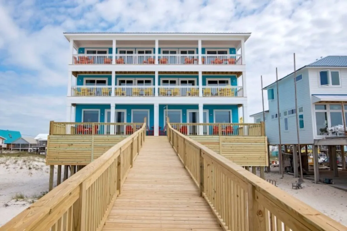The image showcases a stunning three-story beach house painted in soothing blue and white tones, perched on stilts right above the sugar-white sands of Gulf Shores. A spacious wooden walkway leads directly from the pristine beach to the house's entrance, while multiple balconies on each floor promise sweeping ocean views and ample outdoor relaxation space. The tranquil, uncrowded beach setting hints at the peaceful getaway awaiting guests.