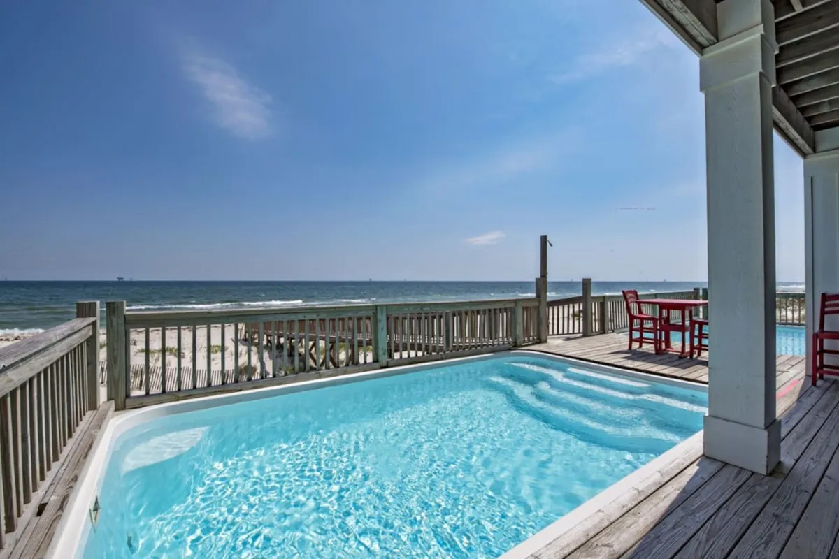 A bright, inviting private swimming pool is the focal point, surrounded by a wooden deck furnished with red seating. The scene highlights the unobstructed Gulf beach and ocean horizon beyond the deck railing, with wooden stairs leading directly to the sand. The tranquil ambiance is enhanced by the open sky and shimmering pool water.