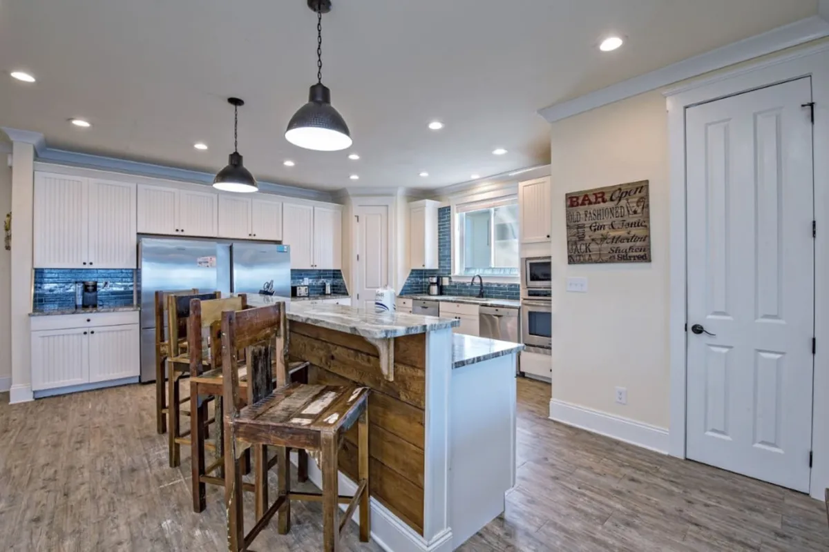 A state-of-the-art kitchen with a marble-topped island and rustic wooden bar stools, complemented by a modern stainless steel refrigerator, white cabinetry, and a blue tile backsplash. The space is bright, inviting, and designed for both meal preparation and casual socializing.