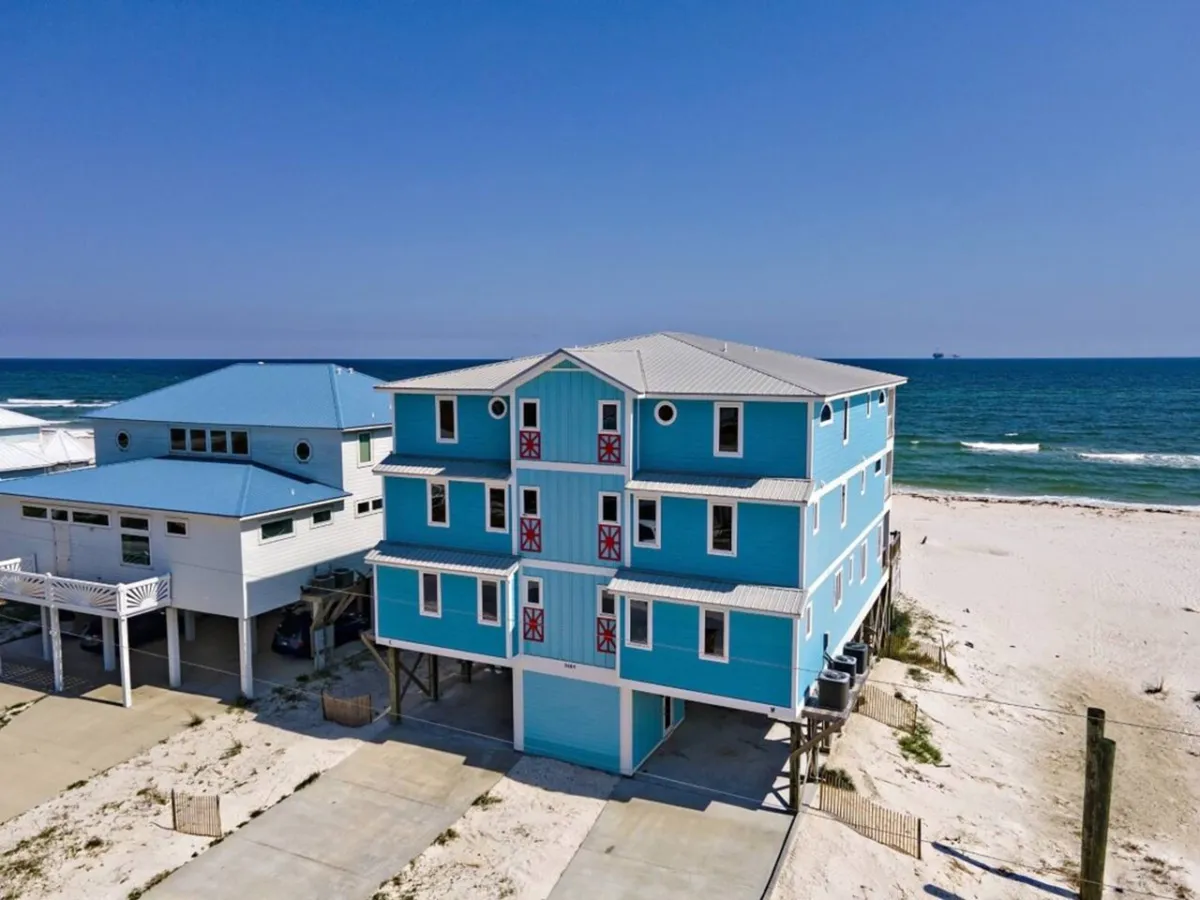 A large blue duplex beach house elevated on stilts, fronted by a wide expanse of sugar-white sandy beach that stretches to the turquoise Gulf of Mexico. The clear, gentle waves and neighboring beach houses set a tranquil coastal scene, with ample space for group activities and exploration. The property’s decks and balconies are visible, inviting thoughts of outdoor gatherings after a day’s adventure.