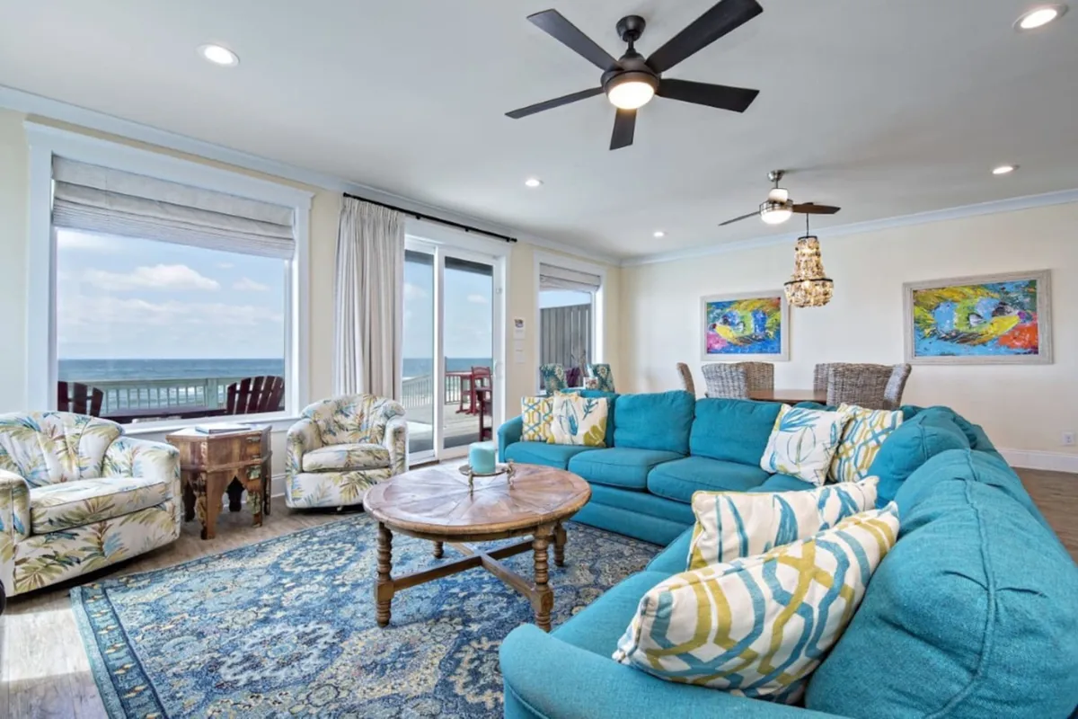 A large turquoise sectional sofa and two floral armchairs are arranged in a cozy seating area facing massive windows with unobstructed ocean views. A round wooden coffee table anchors the space, and the adjacent area leads seamlessly to the outdoor deck.