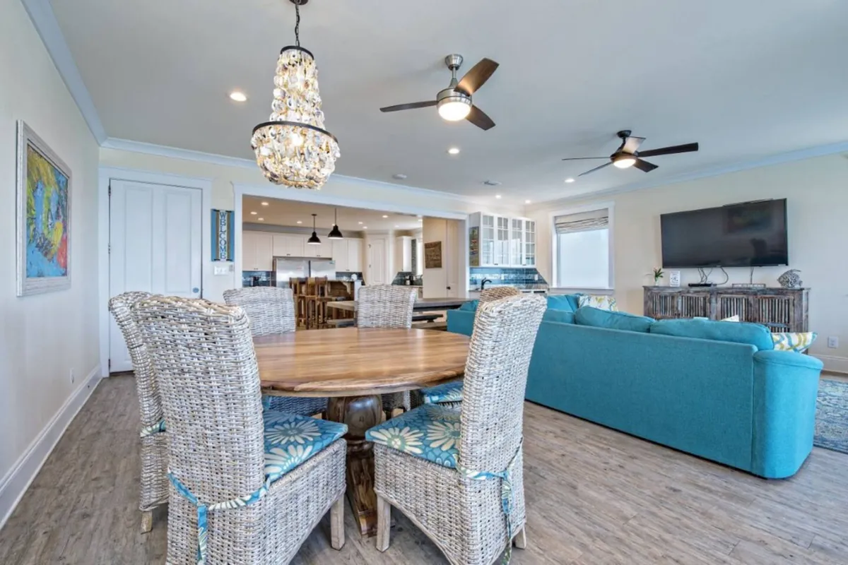 This image displays a bright, open-concept living and dining area with a turquoise sectional sofa, round wooden dining table with wicker chairs, a large flat-screen TV, and a modern kitchen area with appliances. The mood is cheerful and relaxed, evoking a comfortable beach house retreat ideal for group gatherings.