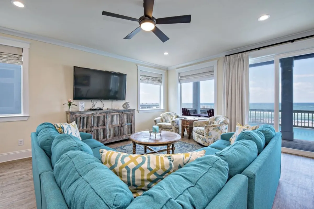 A comfortable interior scene with a large teal sectional sofa, rustic wooden media console with a mounted flat-screen TV, round wooden coffee table, and large windows/sliding doors offering ocean and pool views. The space is sunlit, inviting, and ideal for both relaxation and socializing.