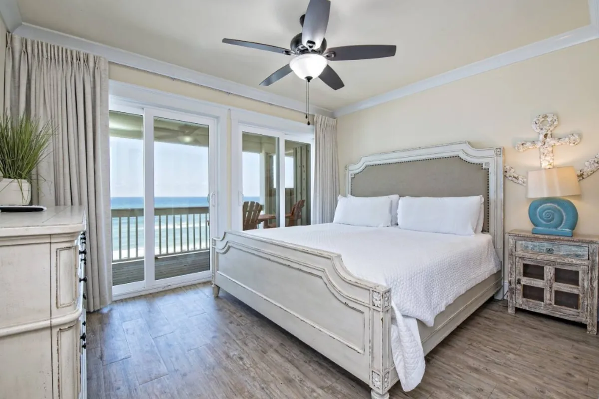 A serene king-sized bedroom with white bedding, large sliding glass doors that open to a balcony with a view of the ocean, a ceiling fan, and a rustic bedside table with nautical decor. The room is filled with natural light and exudes tranquility.
