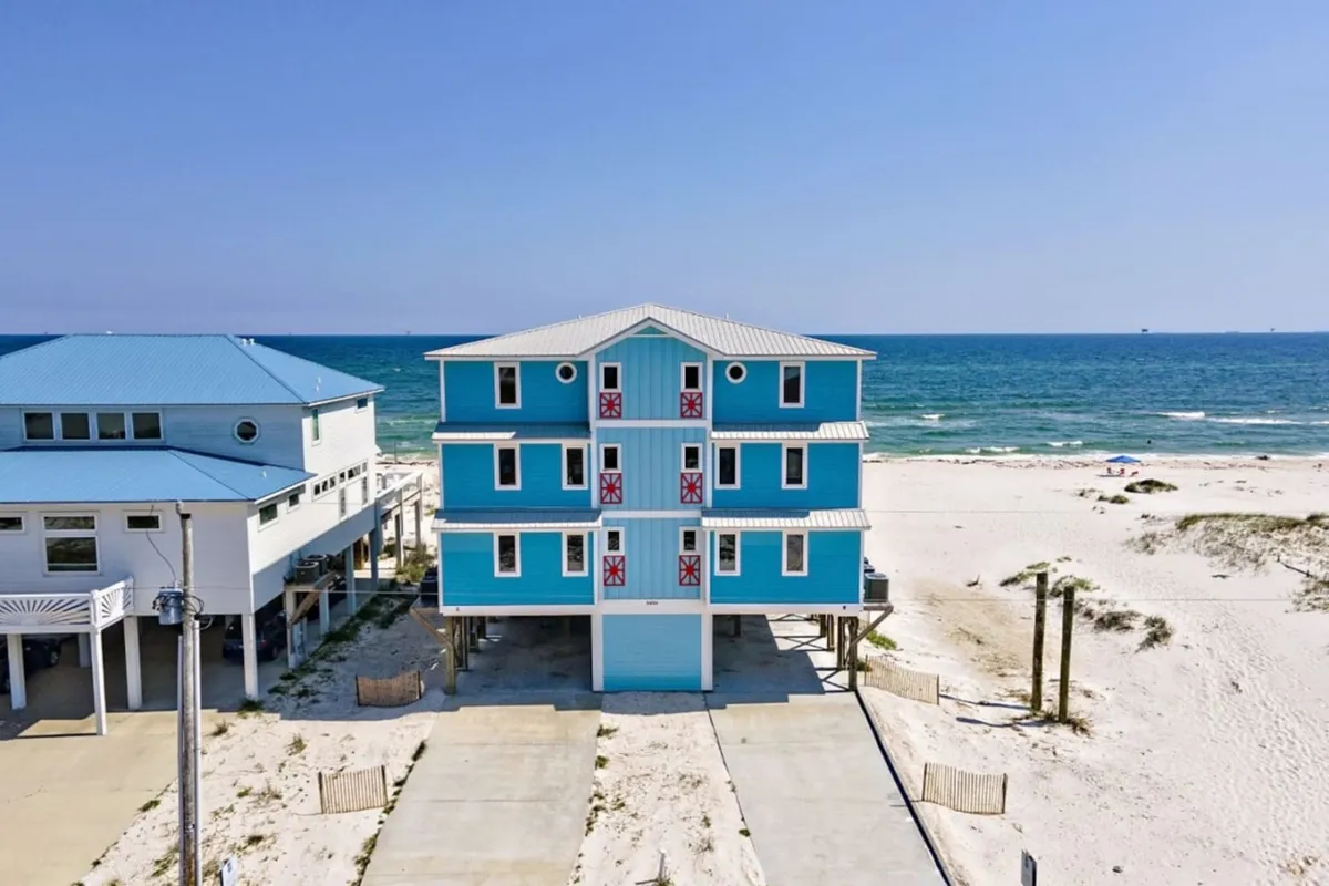 A vibrant blue three-story beach house with red-accented doors and white trim sits atop sugar-white sands, facing the Gulf of Mexico. The scene captures adjacent, similarly styled homes, a clear blue ocean, and gentle waves, creating a sense of exclusivity and relaxation. The home’s balconies and decks are visible, suggesting ample outdoor gathering space.