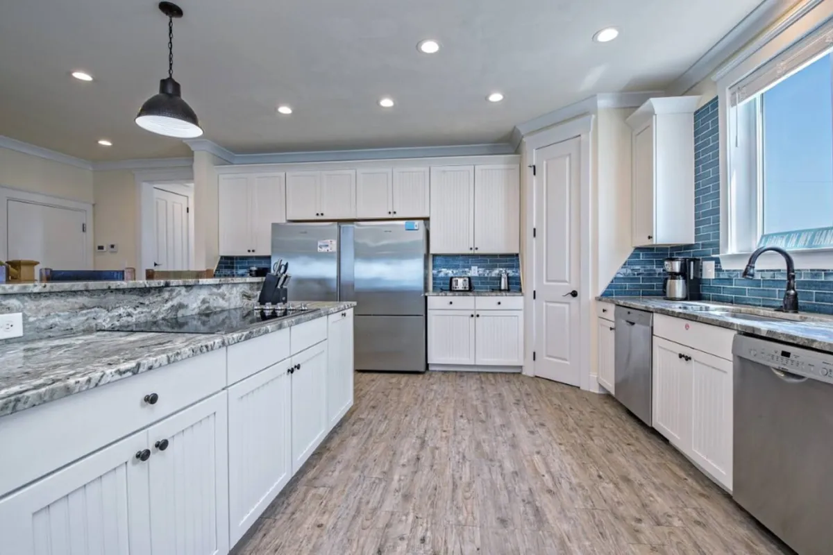 A bright, spacious kitchen with a large granite island, stainless steel refrigerator, blue subway tile backsplash, and a big window that lets in natural light. The island offers ample space for meal prep or group cooking, while the overall layout encourages social interaction and group activities. Coastal touches and an open layout connect the kitchen to the rest of the living space.