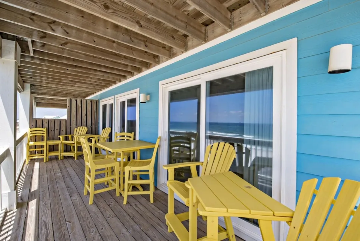 A vibrant exterior deck with yellow Adirondack chairs and tables sits against a blue exterior wall, overlooking the ocean. The wooden deck flooring creates a warm, inviting space, while the sliding glass doors reflect the expansive sea view, suggesting a seamless transition from indoor to outdoor living. The setup is ideal for sunset dining, morning coffee, or casual gatherings, with the ocean visible just beyond the deck.