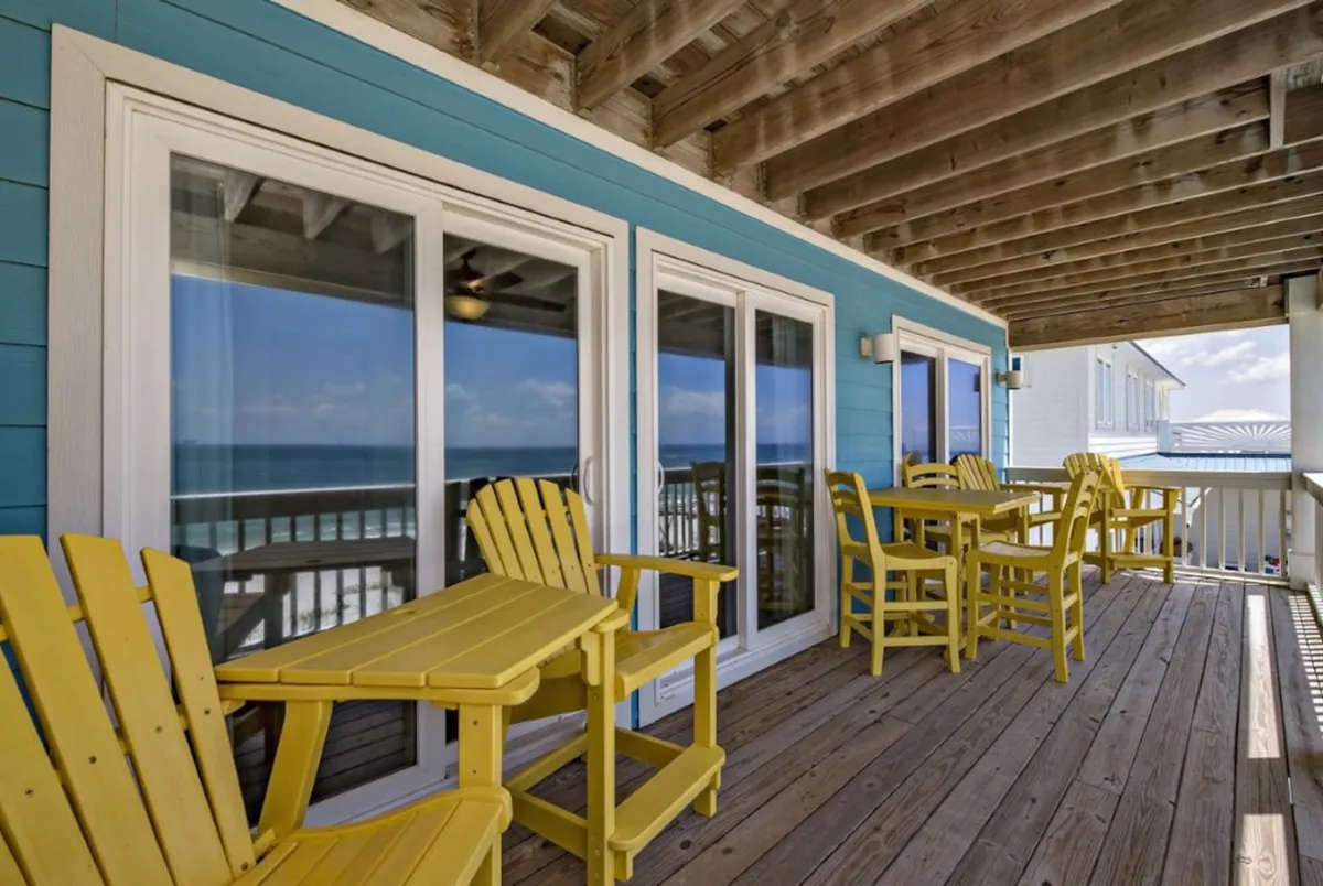 A cheerful porch area features yellow Adirondack chairs and tables set on wooden deck flooring, with a blue painted exterior wall and sliding glass doors. The setup invites relaxation, socializing, and outdoor meals, with the ocean just beyond the deck. The space is casual yet elegant, perfect for enjoying Gulf breezes and sunset views.