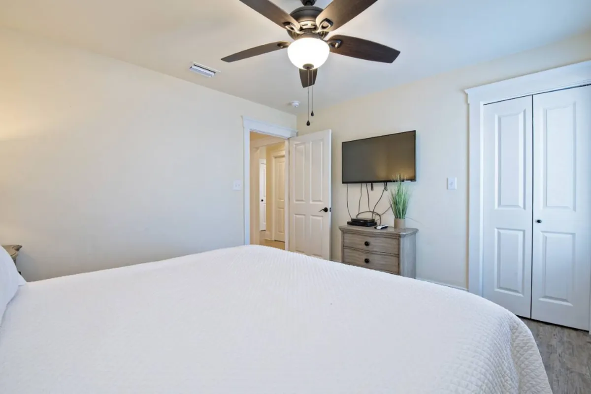 A spacious bedroom with a large bed topped with crisp white bedding, a ceiling fan with light, wall-mounted flat-screen TV, a wooden dresser, and a decorative plant. The setting is peaceful and private.