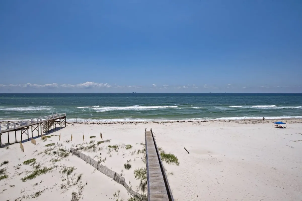 A tranquil walkway leads over pristine dunes to an unspoiled stretch of sugar-white beach. Gentle Gulf waves lap the shore, and a shaded umbrella with a beach chair invites moments of peaceful solitude. The absence of crowds enhances the sense of a secluded oasis.