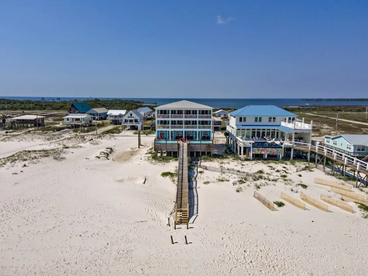 A central beach house with multiple balconies and a long private wooden walkway leading to an expansive white sandy beach. Neighboring elevated beach homes are visible, and the scene conveys spaciousness, privacy, and luxury. The setting is tranquil, with the Gulf of Mexico stretching out in the background.