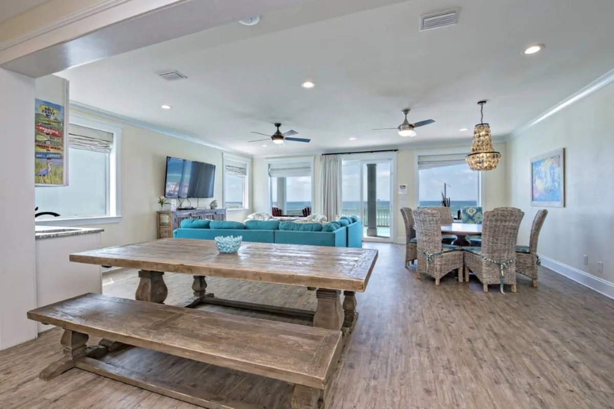 This image captures a spacious, open-plan living and dining area, featuring a large rustic wooden dining table with benches, a turquoise sectional sofa facing a wall-mounted TV, a round dining table with wicker chairs, and expansive glass sliding doors that open to an ocean view. The space is filled with natural light and offers seamless access to the deck outside.
