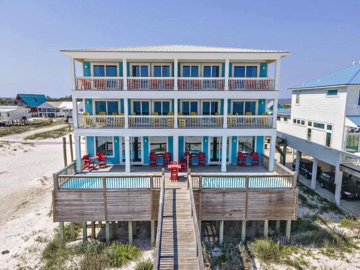 A vibrant three-story beach house with wide balconies overlooking private swimming pools on a ground-level deck. Red Adirondack chairs cluster around the pools, and a boardwalk leads directly from the house to the secluded sandy beach, highlighting the seamless blend of indoor and outdoor group spaces.