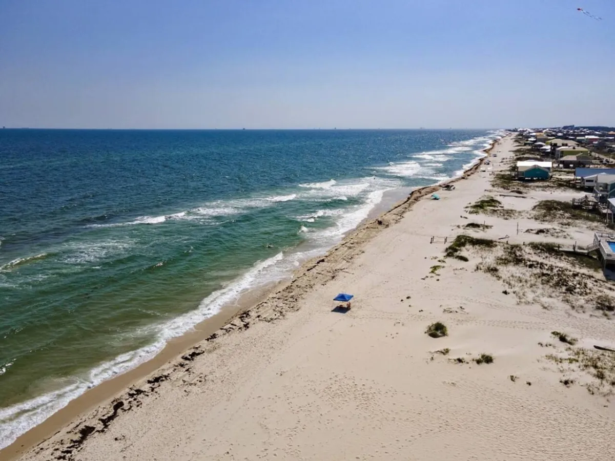 A tranquil scene of a sugar-white sandy beach bordered by calm Gulf waters, with a single blue beach umbrella and a line of beachfront houses in the distance. The beach is nearly empty, emphasizing peace, solitude, and a private atmosphere.