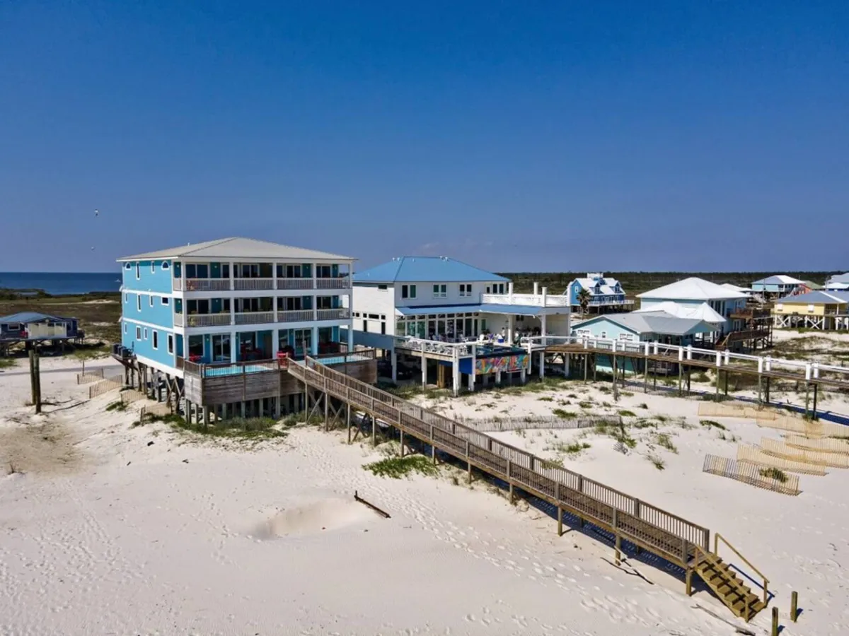 A large, elegant blue three-story beach house on stilts with a wooden boardwalk extending from the home to the sugar-white sandy beach, with clear turquoise Gulf waters and neighboring homes set apart by natural dunes and greenery.