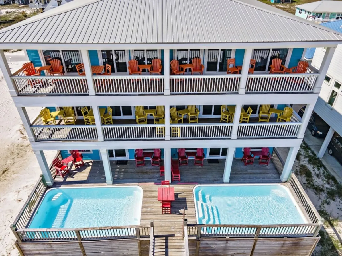 The image shows the exterior of the duplex property, featuring two private swimming pools, three levels of balconies with colorful Adirondack chairs, and a wooden deck area between the pools. The symmetrical design highlights the separation between the two sides of the property and the private amenities available to each group.