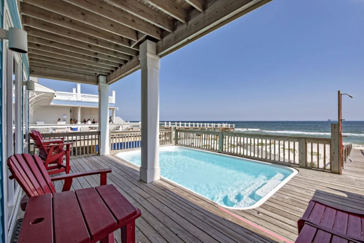A serene outdoor deck featuring a private swimming pool with an ocean view, red Adirondack chairs for lounging, and a wooden pier extending into the water. The pool area is calm and inviting, just steps from the surf, offering both relaxation and spectacular views of the Gulf.