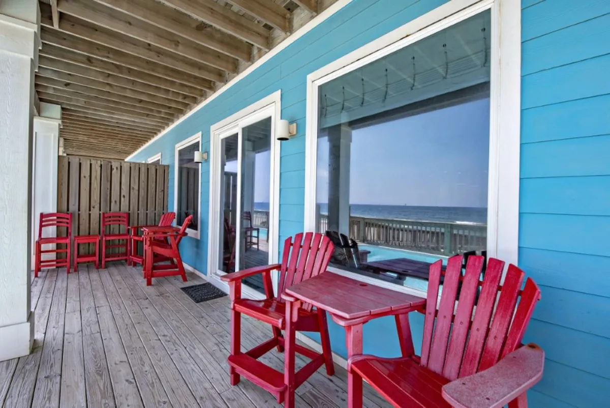A cheerful cluster of bright red Adirondack chairs and tables sits atop a wooden deck, framed by a blue exterior wall with white trim. The reflection of the ocean in the windows hints at the proximity to the water, while the open seating invites social gatherings and relaxation. The overall mood is welcoming and sunlit.
